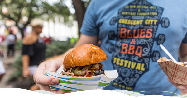 Person holding a barbecue sandwich wearing a shirt with a guitar logo of the Crescent City Blues & BBQ Festival at the Crescent City Blues & BBQ Festival.