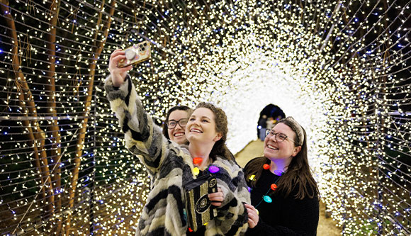 Three friends take a selfie while standing in a tunnel of sparkling holiday lights.