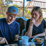 Two people eat dinner at a riverfront restaurant. Behind them is a bridge.