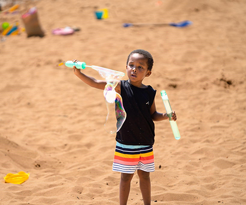 A picture of a boy playing with his bubble maker on a sandy beach.