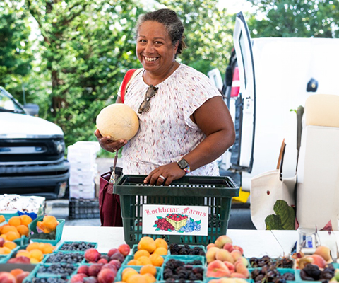 Picture of a woman at a farmers market stand holding a cantaloupe while shopping.