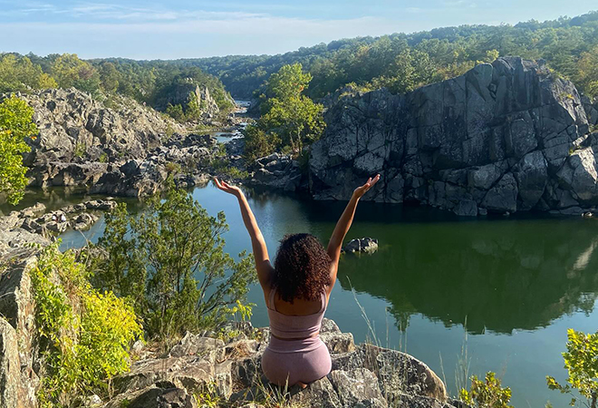 Picture of a woman sitting on top of a mountain point overlooking the water and mountains.
