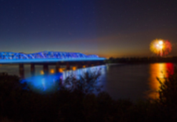 The Harahan Bridge at night illuminated in blue over the Mississippi River with fireworks in the background.