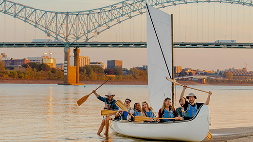 A group paddling to shore in a canoe on the Mississippi River with the Hernando de Soto Bridge in the background.