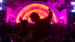 A couple with their arms raised while looking at each other during at show at Overton Park Shell. In the background, a crowd gathers at the stage which is lit with orange and pink lights.