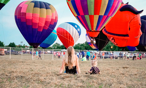 A woman and child sit on the ground watching several colorful hot air balloons take flight.