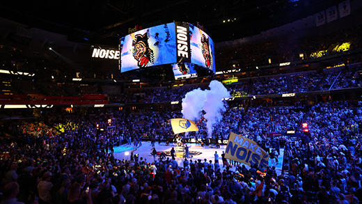 Inside the FedExForum with the lights lowered and Memphis Grizzlies flags waving on the court as the crowd is on their feet. The Grizzlies logo is displayed on the jumbotron while a smoke machine bursts puffs of smoke in the background.