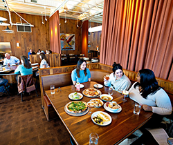 Three women with a table full of pizza, cocktails and other dishes, with other diners in the background, at Hog and Hominy.