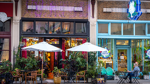 An exterior of Juiced What You Needed juice cafe on South Main St., with plants and patio furniture out front and purple string lights above the entry. A woman sits next door at a patio table outside neighboring The Book Juggler book story.