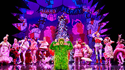 Actors on stage for Dr. Seuss' How the Grinch Stole Christmas! The Musical with a Christmas tree in the background and a set design showing presents. 
