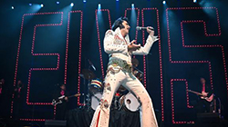 An Elvis tribute artist performing with a band on stage with a neon red ELVIS sign behind him.