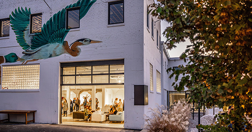 An exterior view of Ugly Art Co., with people gathered inside the gallery space and a mural of a bird above the entrance.