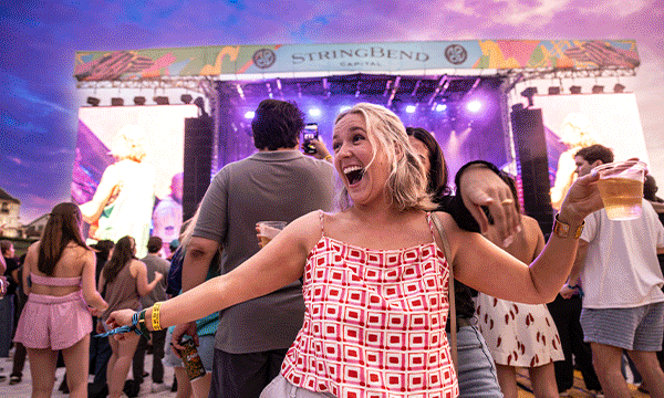 A rotating series of images from Memphis' RiverBeat music festival, including a woman dancing with her back turned to the crowd and stage, a large crowd gathered at a stage hitting inflatable beach balls into the air, an aerial view of a crowd gathered at a stage at the festival with the Mississippi River, the Hernando de Soto Bridge and the Memphis skyline in the background, and a stage at night illuminated in orange and red lights with a crowd in front with their hands up. 