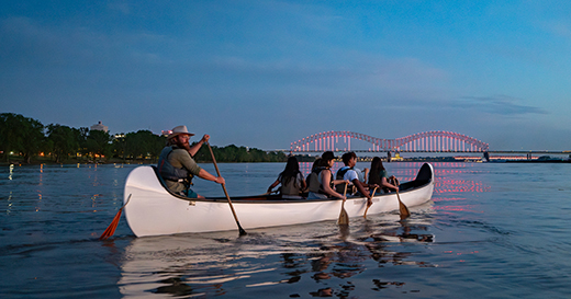 A group on a guided canoe trip with Mississippi River Expeditions on the river at dusk with the Hernando de Soto Bridge aglow in pink neon lights in the background.