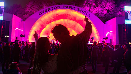 A couple with their arms raised behind a crowd of people enjoying a concert at Overton Park Shell, with the shell in the background illuminated in pink and orange neon lights.
