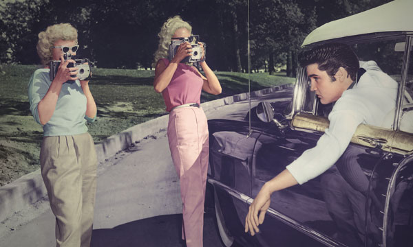 A photo of Elvis leaning out of the driver's seat of his car as he passes two women taking pictures of him. 