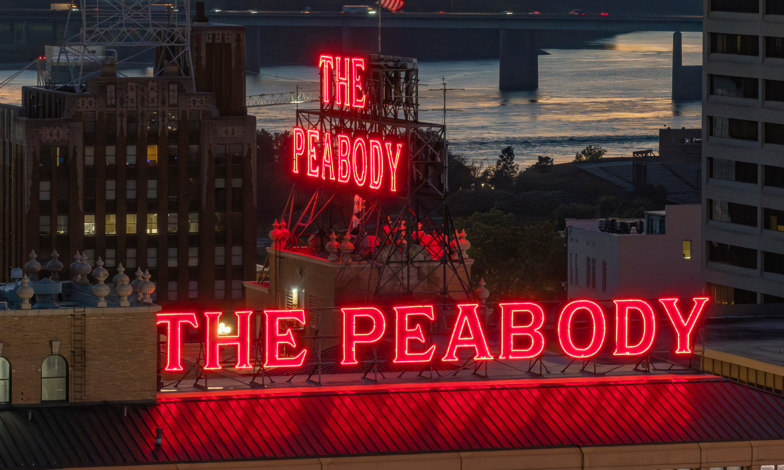 An aerial view of the neon red 'The Peabody' signs with buildings and the Mississippi River in the background.