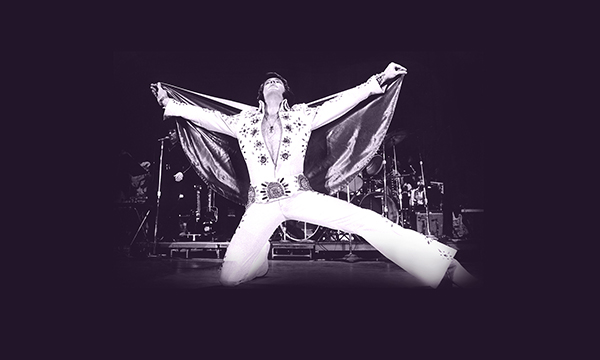 An image of Elvis on one knee on stage, while wearing one of his iconic jumpsuits. His arms are outstretched, holding the cape of his stage costume.