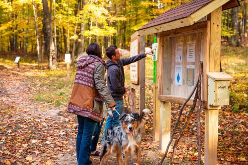 A couple with a leashed dog at a trailhead sign in the woods. 