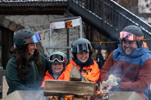 A family in ski gear sitting around a fire pit in the snow. 