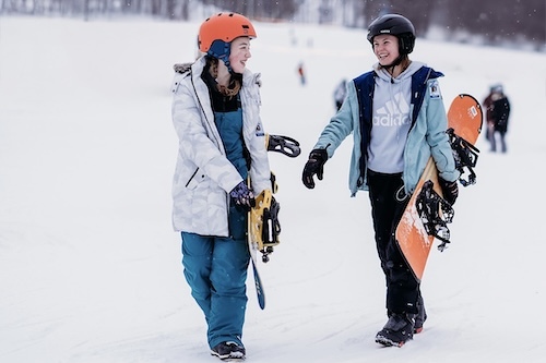 Two women walking with snowboards in the snow. 