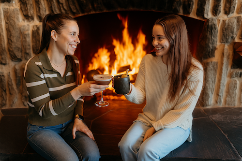 Two women toasting with cocktails by a fireplace. 