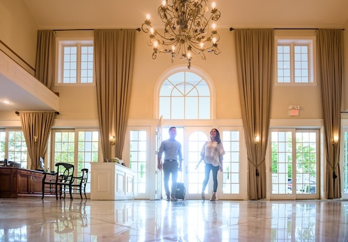 couple coming into the winery inn rolling their suitcase behind them.  the sun is glaring through the glass pane in the door.