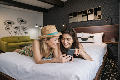 two women laying on the bed looking on their phone and smiling.