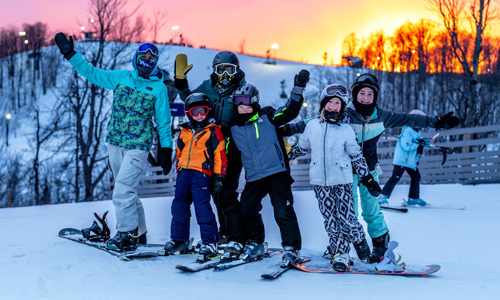 group of skiers waving to the camera as the sun is setting behind them at crystal mountain ski lodge.