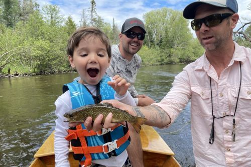 A man holding a small trout beside a boy in a canoe while fishing in a river. 