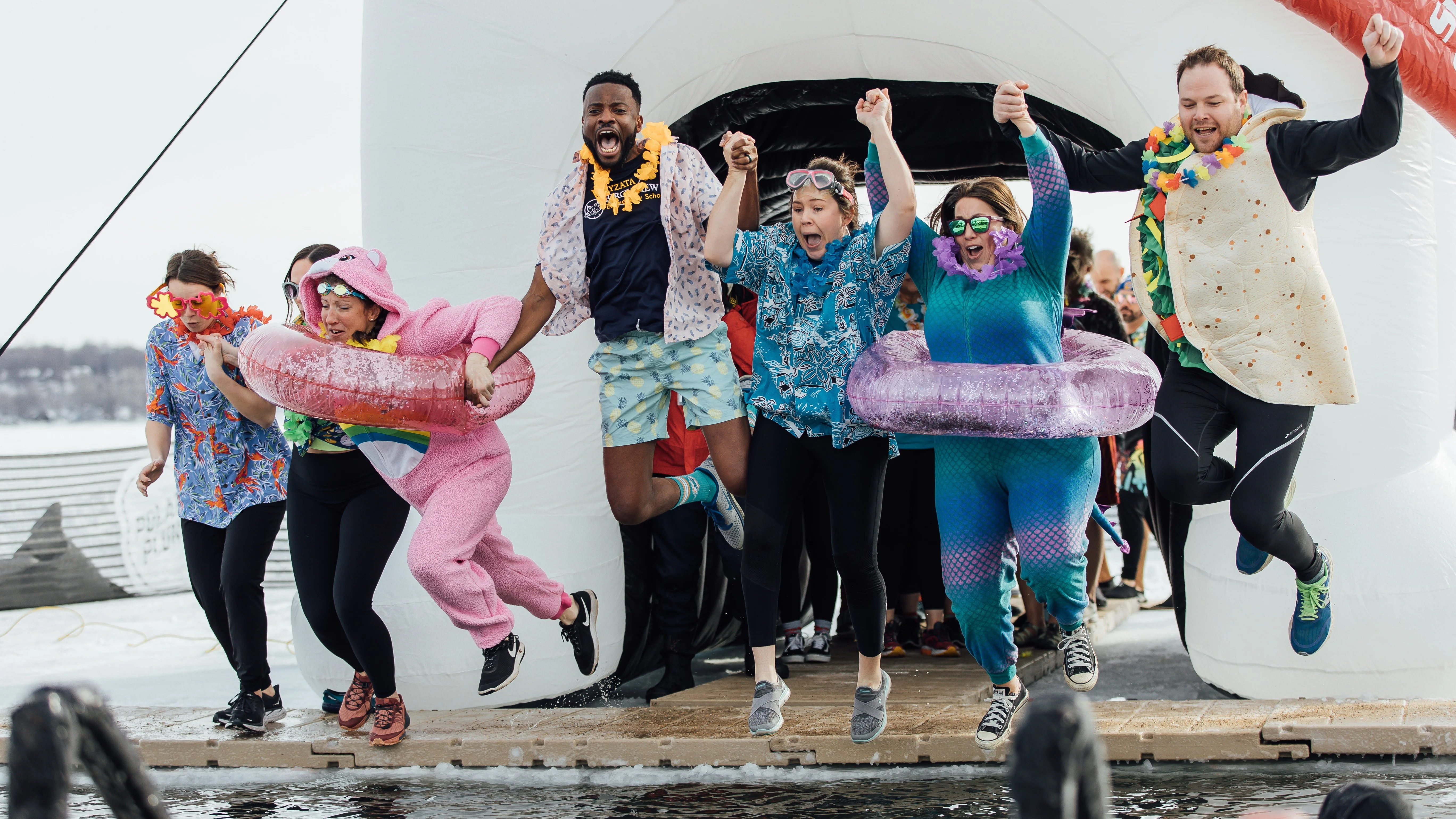 seven people in various fun costumes hold hands and jump into an icy cold lake