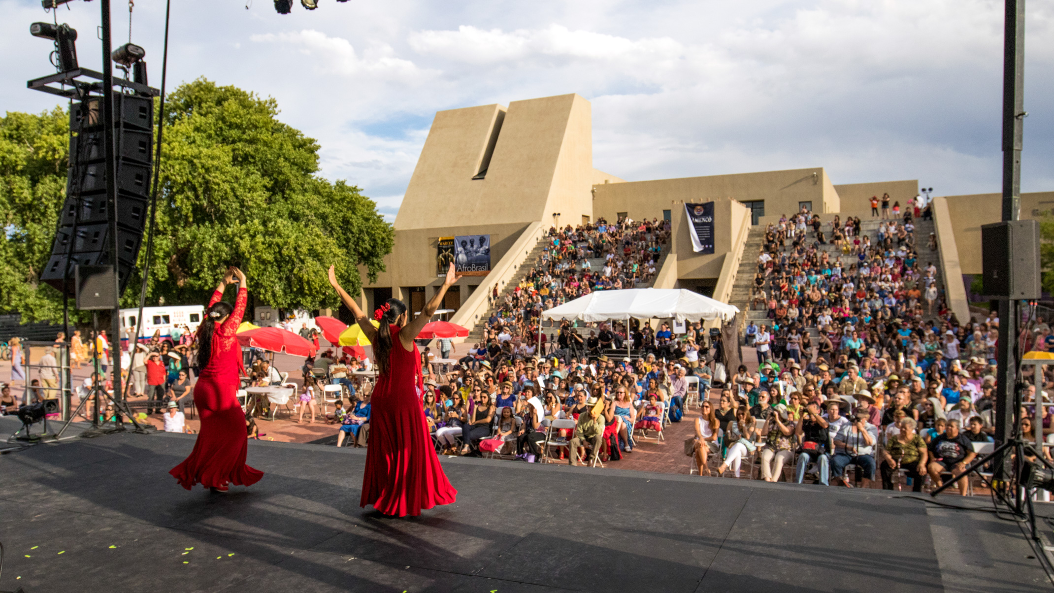 Two flamenco dancers in red dresses perform on an outdoor stage in front of a large crowd seated in an amphitheater on a sunny day.