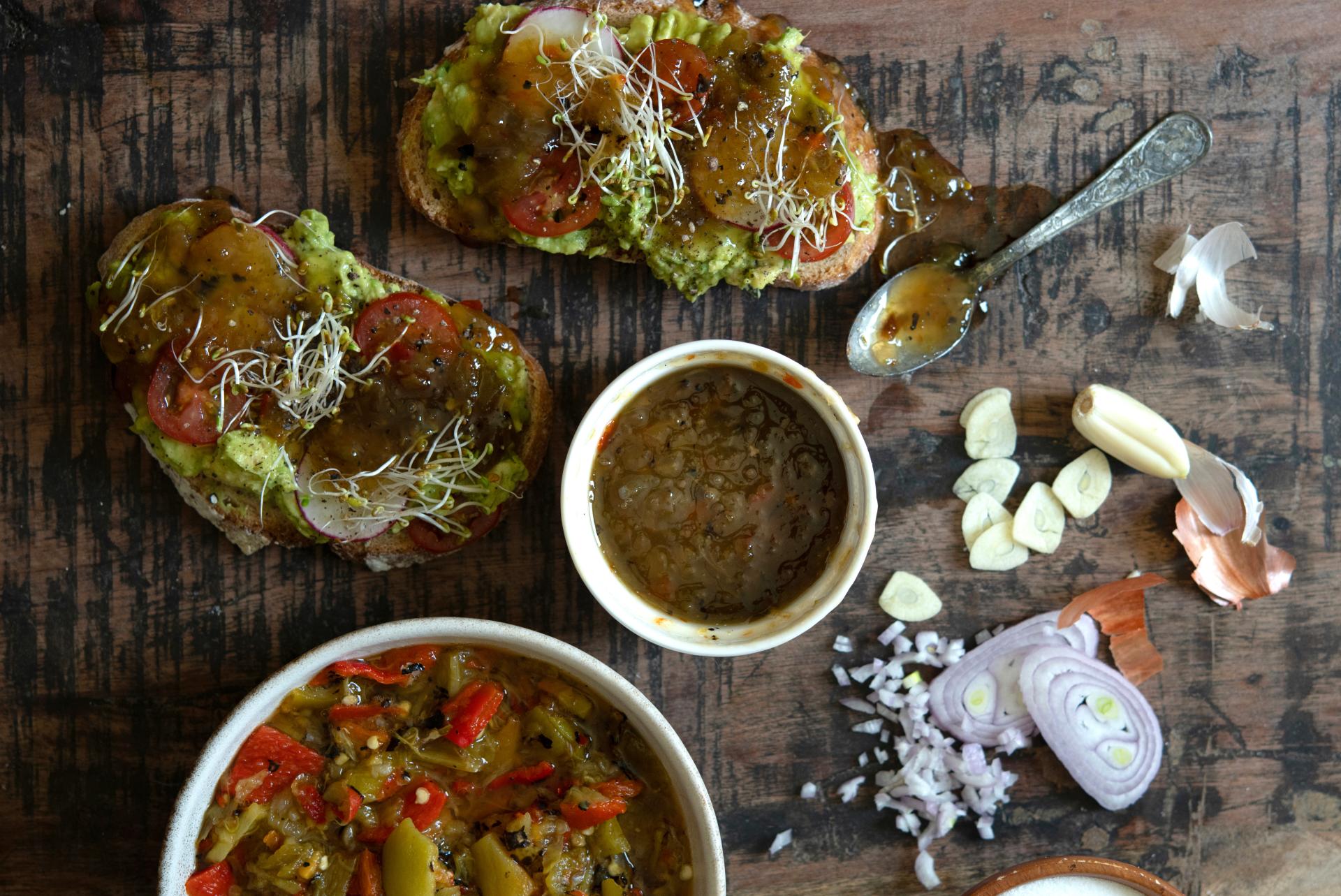 Overhead view of a rustic wooden table with various dishes including avocado toast topped with tomatoes and chile and sprouts, bowls of salsa, and a mix of chopped onions and garlic spread around.