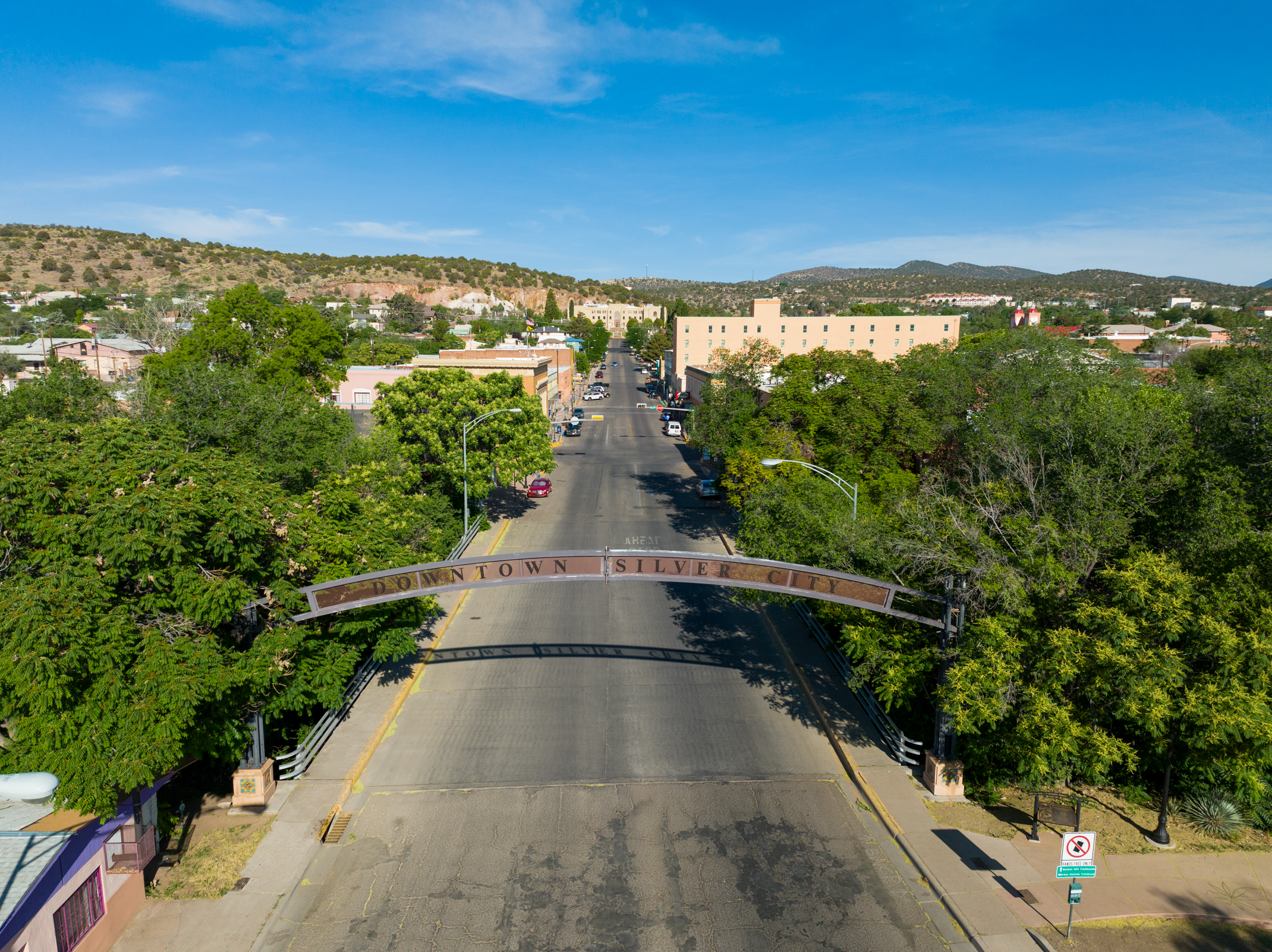 Aerial view of a street entering downtown Silver City, with a prominent arch bearing the city's name over the road, surrounded by green trees and buildings under a clear blue sky.