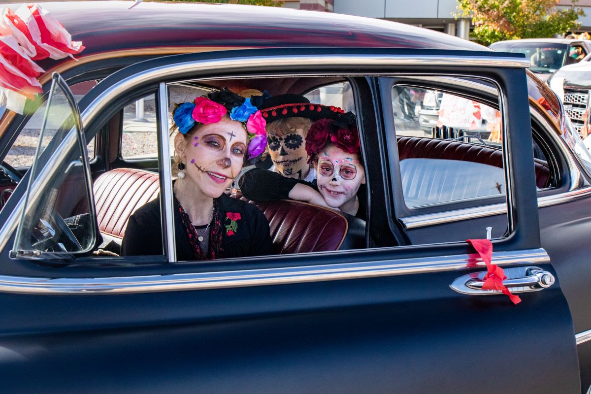 Three people in Day of the Dead makeup sitting in a classic car, smiling at the camera.