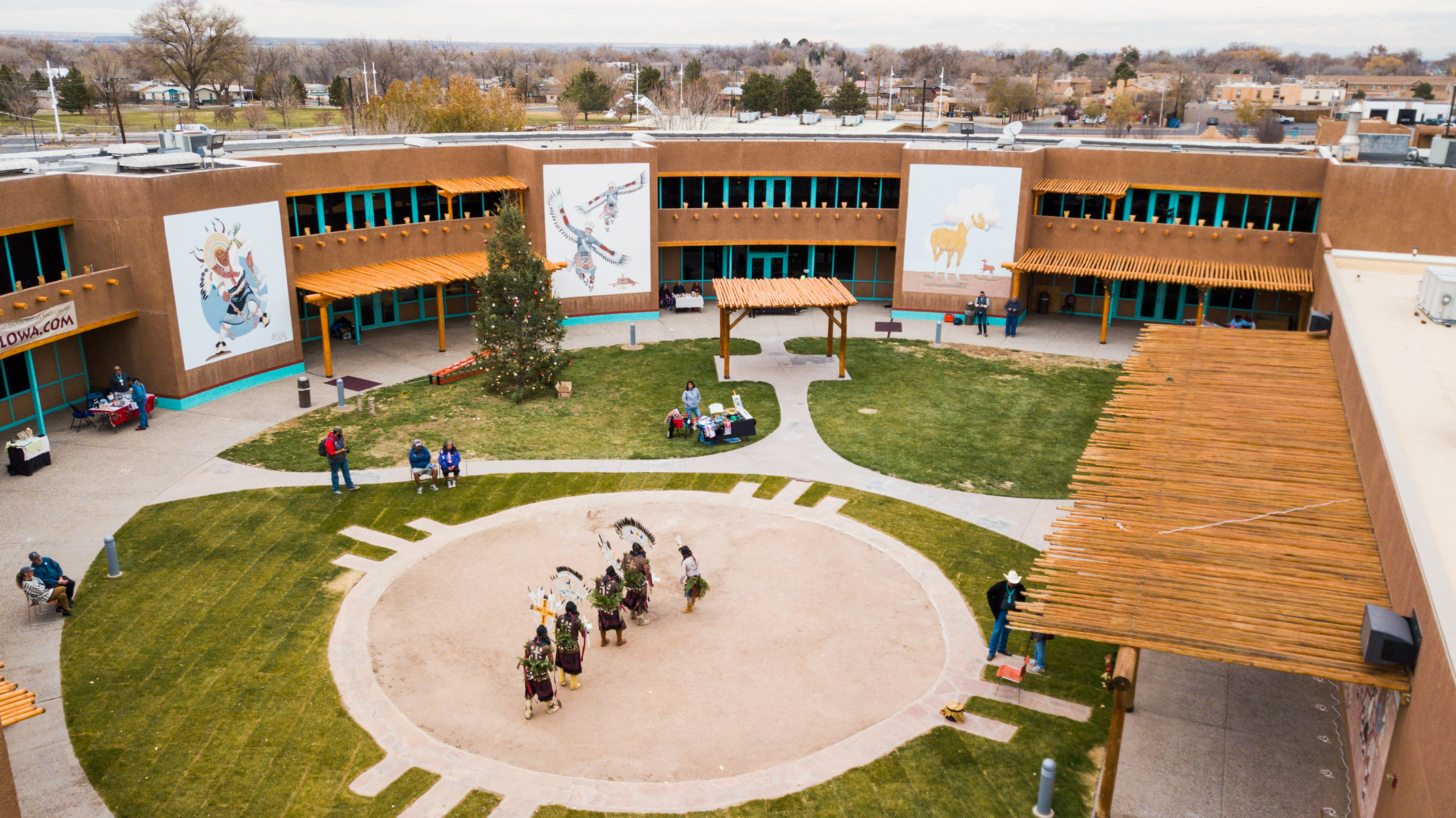 Aerial view of a courtyard at the Indian Pueblo Cultural Center, featuring people gathered around a traditional dance performance, surrounded by buildings with murals and southwestern architecture.
