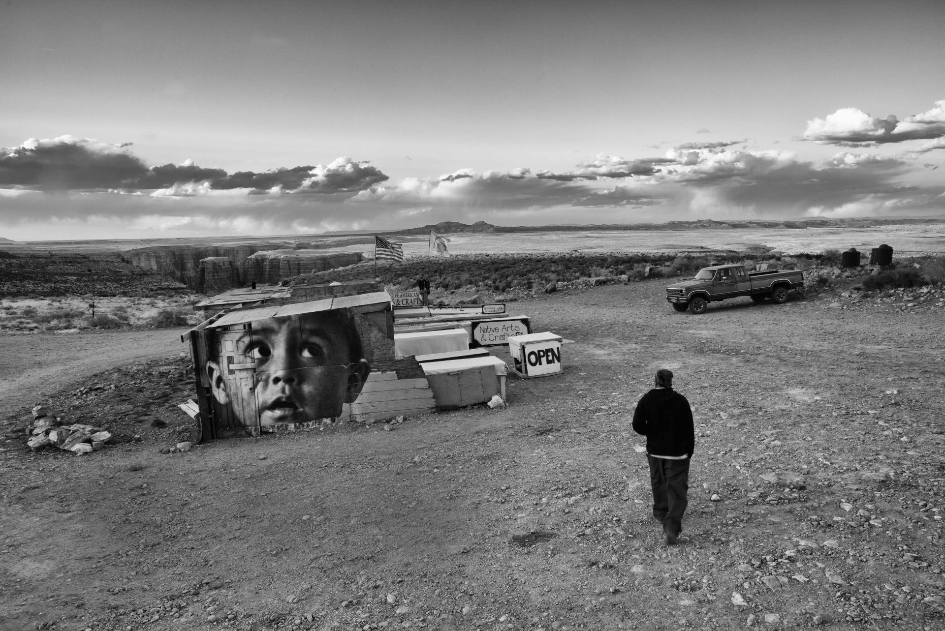 A black and white photograph depicts a person walking towards a small, rustic building adorned with a large mural of a child's face, in a barren desert landscape. Nearby, there is an "OPEN" sign and a parked car. Distant mountains and expansive skies are visible in the background.