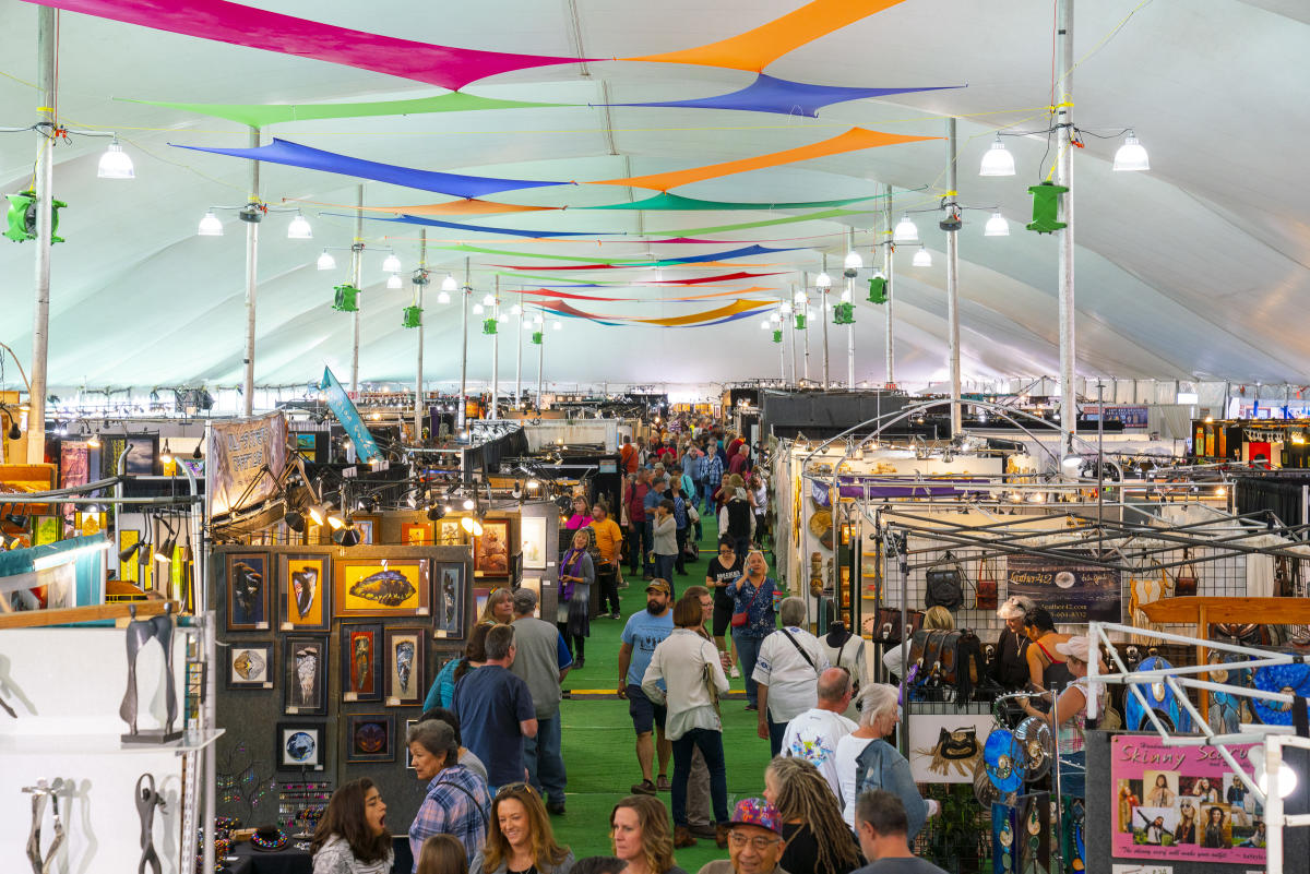 patrons walk between booths featuring arts and crafts in a large room with lights and colorful streamers hanging from rafters