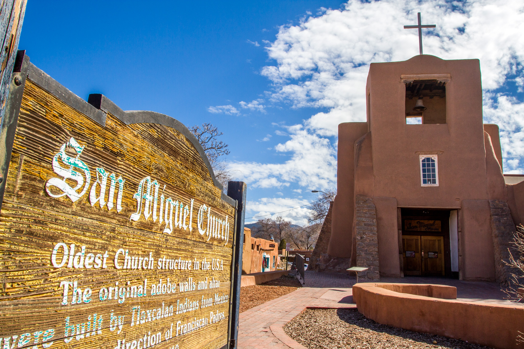 The San Miguel Chapel, known as the oldest church structure in the U.S., with adobe walls and a wooden sign in the foreground detailing its history, located under a clear blue sky.