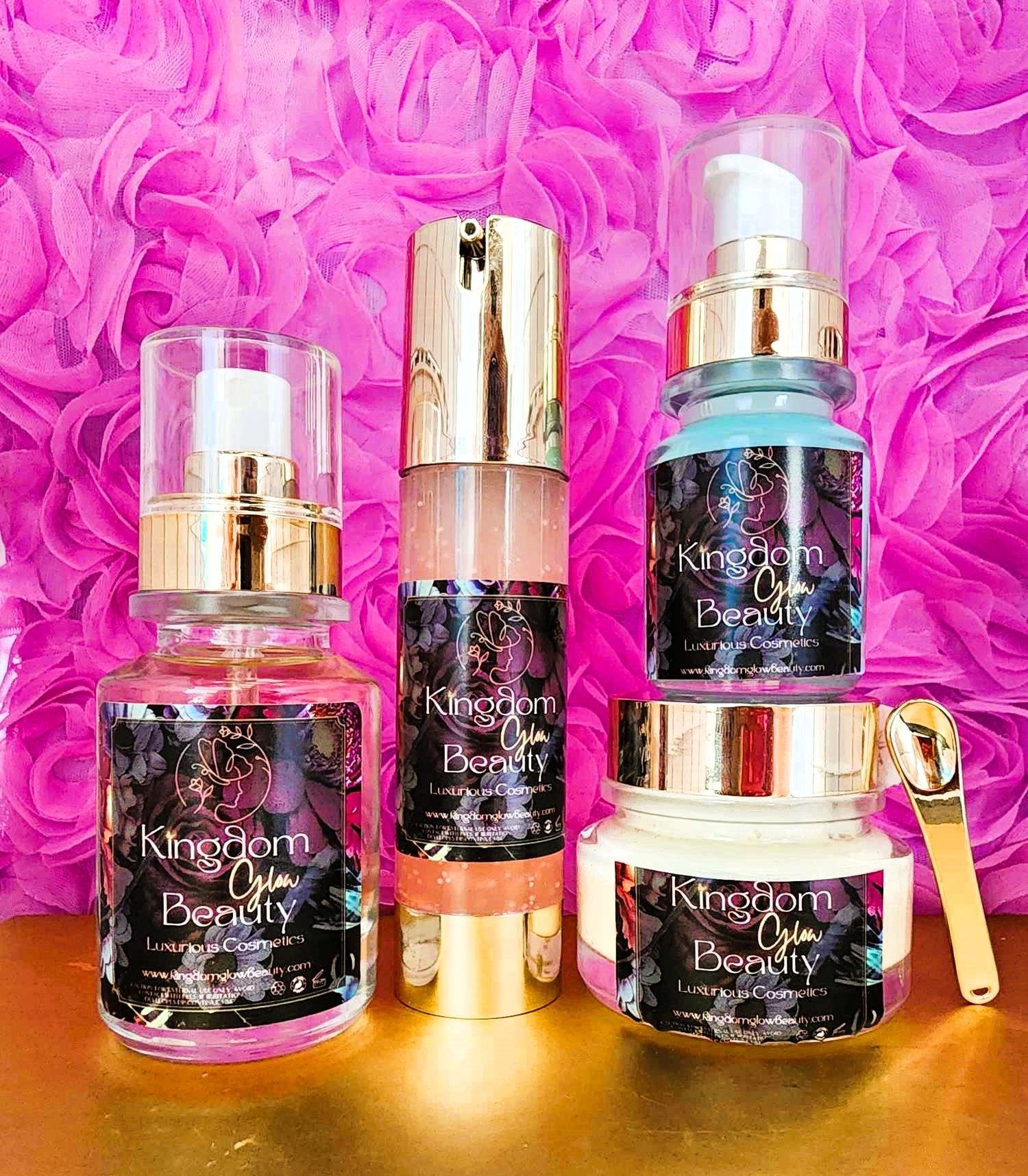Four Kingdom Glow Beauty skincare products arranged against a vibrant pink floral background. From left to right: a glass bottle, a pump dispenser, another taller pump dispenser, and a jar with a golden spoon on top. All items feature elegant labels.