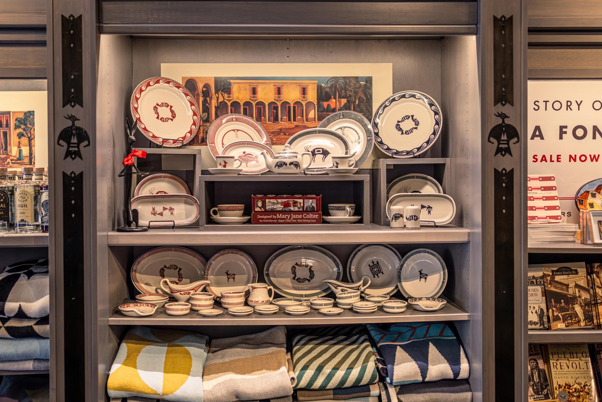 A display shelf with a variety of ceramics, including plates and bowls, featuring historical and architectural designs. The collection is neatly arranged inside a store with promotional posters visible.