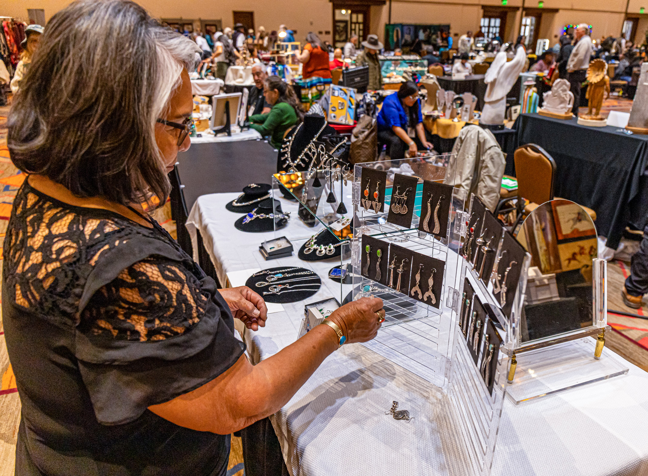 A person examines some jewelry at an indoor market scene with various stalls displaying arts and crafts, attended by people browsing items. Signage indicates this event is related to Santa Fe's Indian Market, a notable annual event.