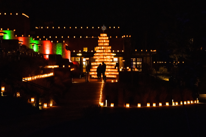 Nighttime view of a Christmas Tree constructed of farolitos or luminarias, featuring a silhouette of a person walking towards the building.
