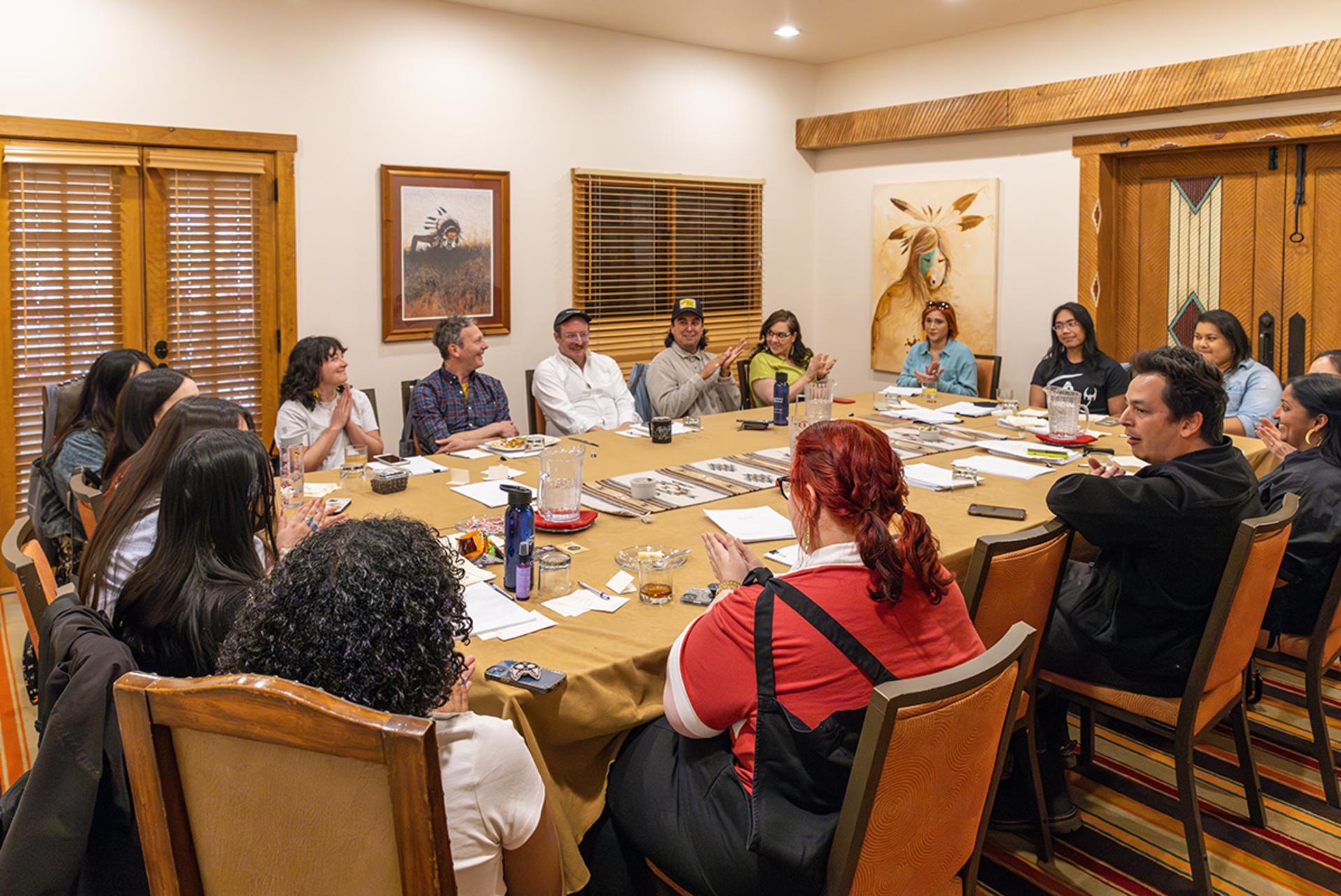A group of people seated around a conference table engaged in a discussion. The room features warm lighting and is decorated with framed artwork, including an image of an indigenous person