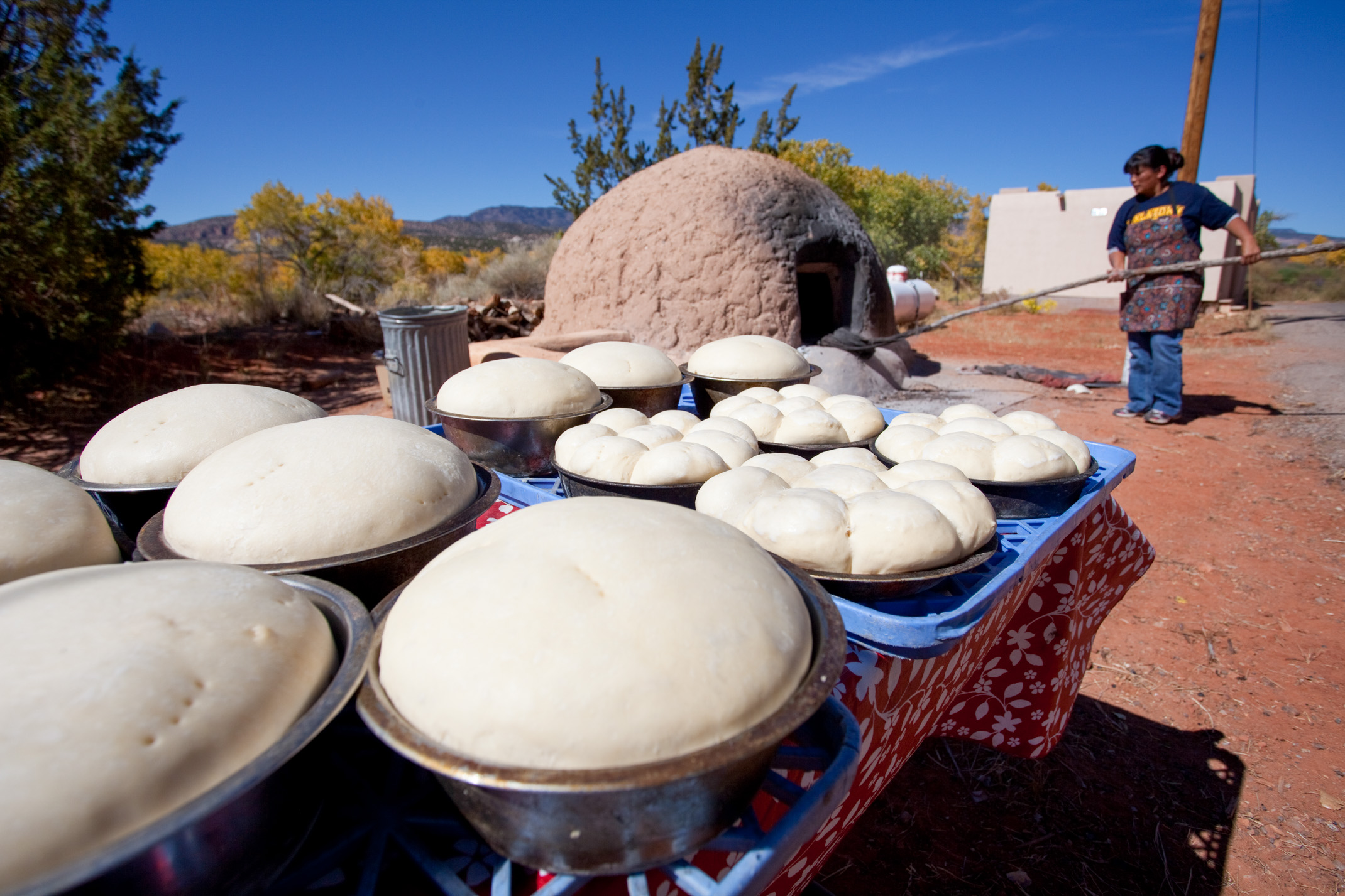 Rising trays of dough sit in pans on a table with a person feeding dough into a traditional clay cooking device with a long stick in the background