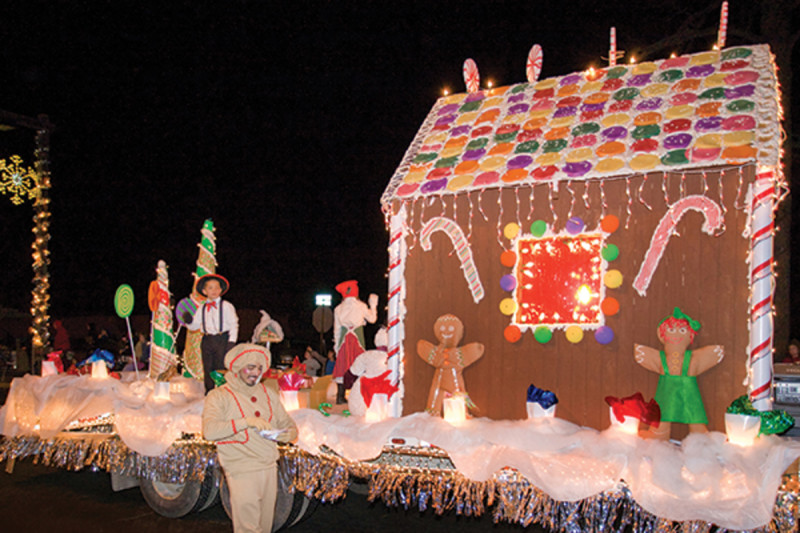 A festive parade float decorated to look like a giant gingerbread house, complete with candy decorations and glowing lights. Several people in holiday-themed costumes, including a gingerbread person and an individual dressed in a white outfit with a green sash, are accompanying the float. The scene is illuminated and cheerful, captured during a nighttime event.