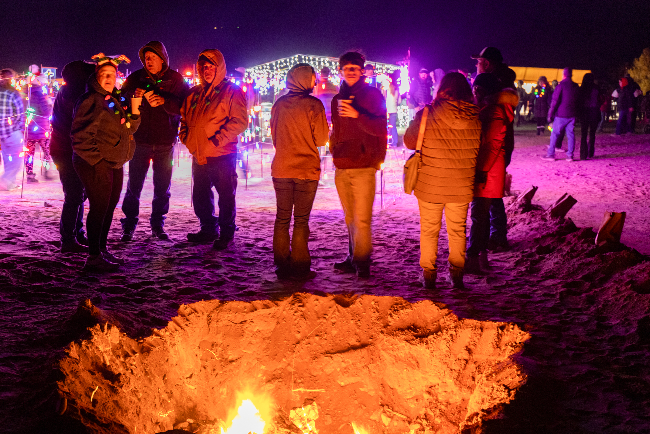 Group of people standing around a bonfire on a beach at night, with illuminated festive lights in the background.