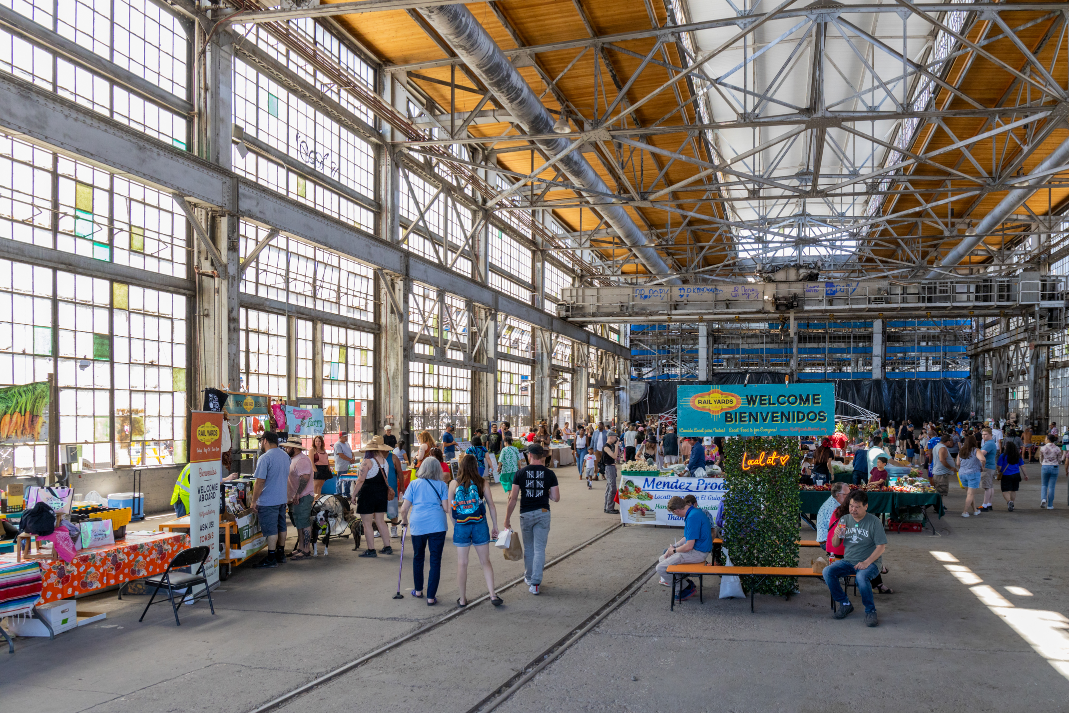 Indoor market at the Rail Yards in Albuquerque, with multiple vendors and visitors around stalls selling various goods. A welcome sign is prominently displayed.