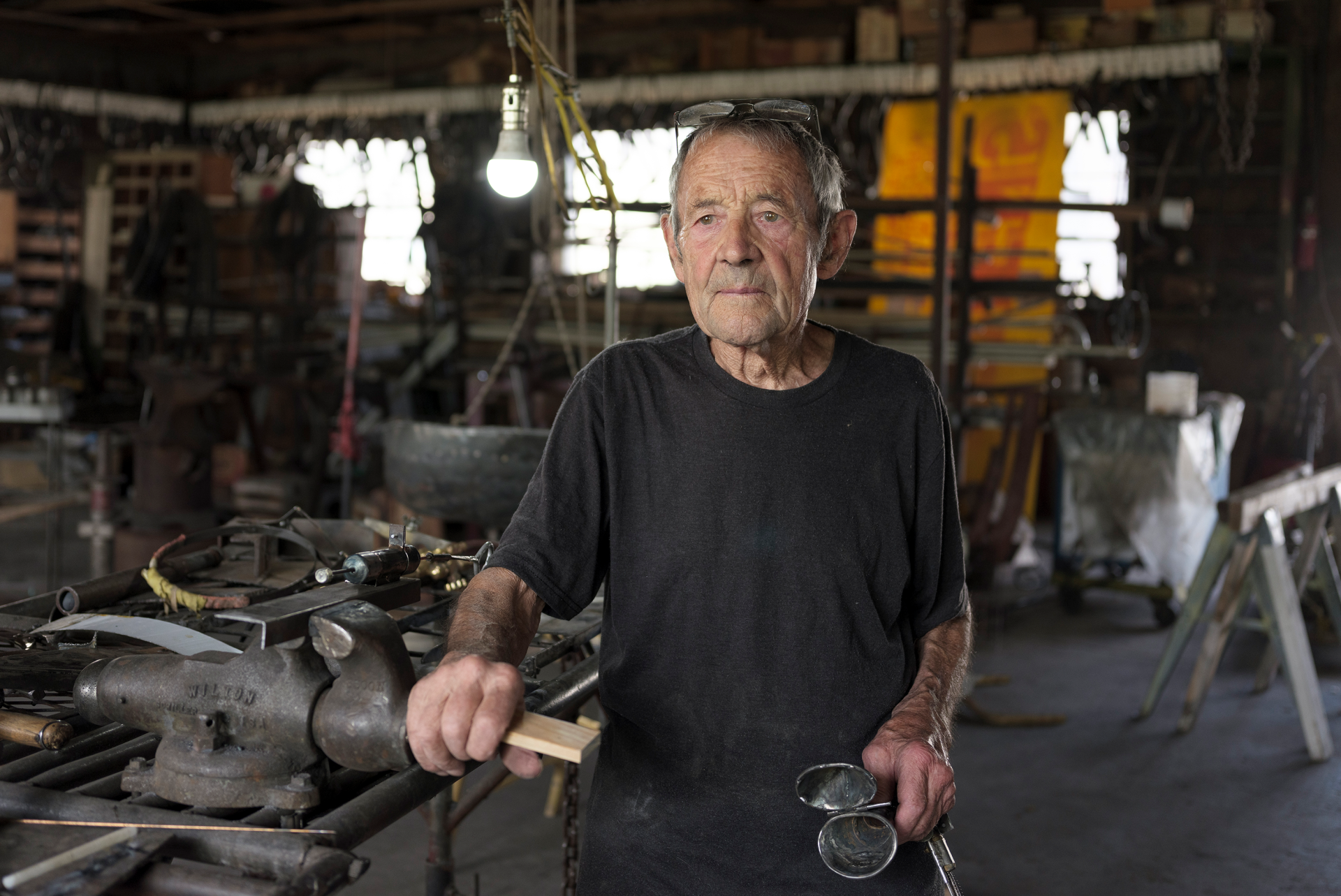 An elderly craftsperson stands in a workshop surrounded by tools, holding a hammer and wearing goggles around their neck.