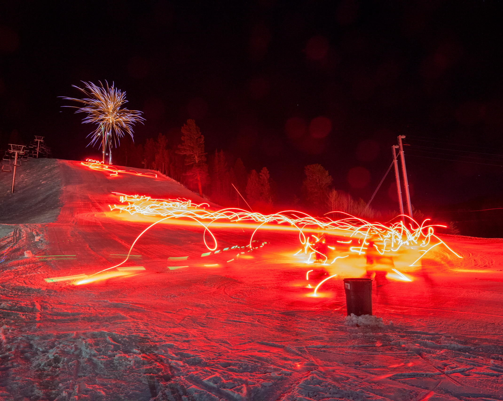 Fireworks illuminate the night sky above a snow-covered ski slope, where a group of skiers holding bright red torches create light trails as they descend.